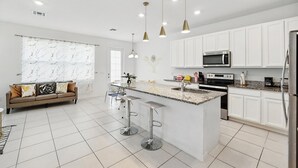Uma cozinha moderna com armários brancos, uma ilha central com banquetas, eletrodomésticos de aço inoxidável e iluminação pendente.