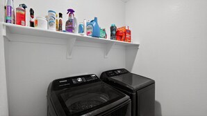 A laundry area with two washing machines and a shelf above containing various cleaning products. - Arizona Vacation Home 3 Bedroom Home (Maricopa)
