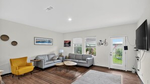 A modern living room with a sofa, armchair, and coffee table, a flat-screen TV mounted on the wall, and a door leading to a balcony. - Arizona Vacation Home 3 Bedroom Home (Maricopa)