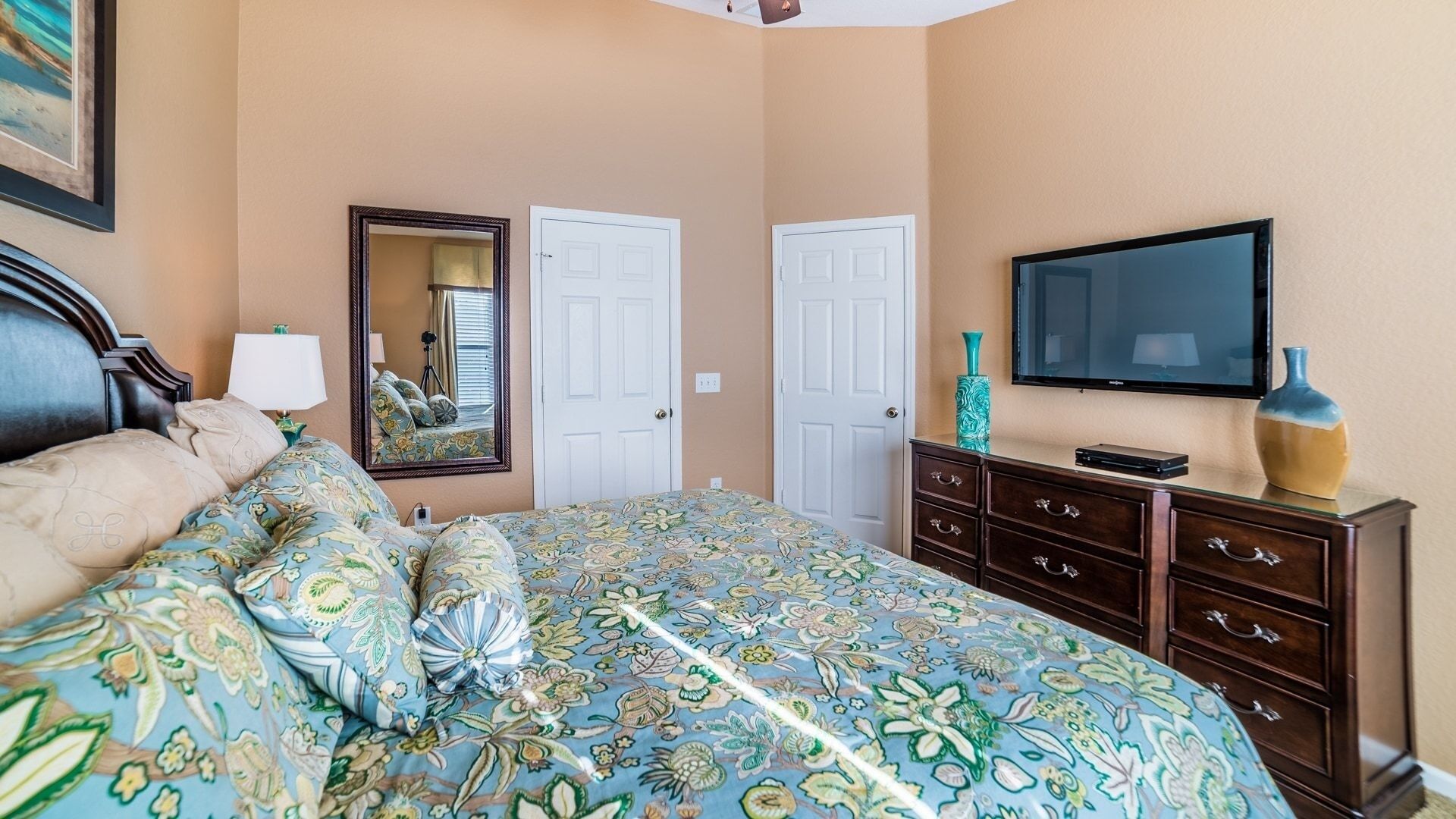 A bedroom with a floral bedspread, a wooden dresser, a television, and a mirror.