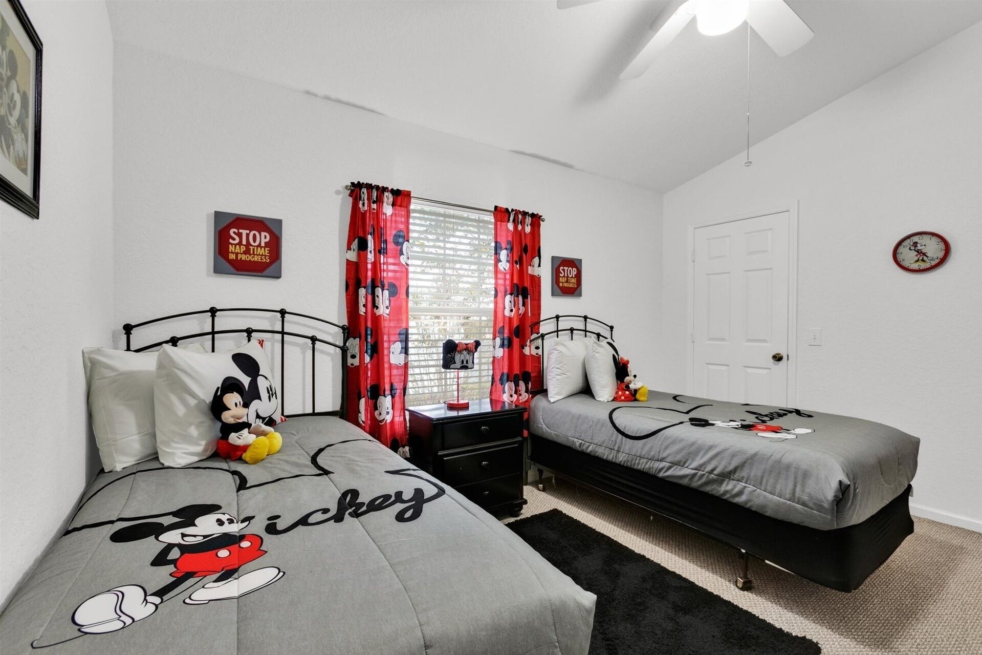 A bedroom with two beds, each with Mickey Mouse-themed bedding. There are two nightstands, a window with red curtains, and a door.