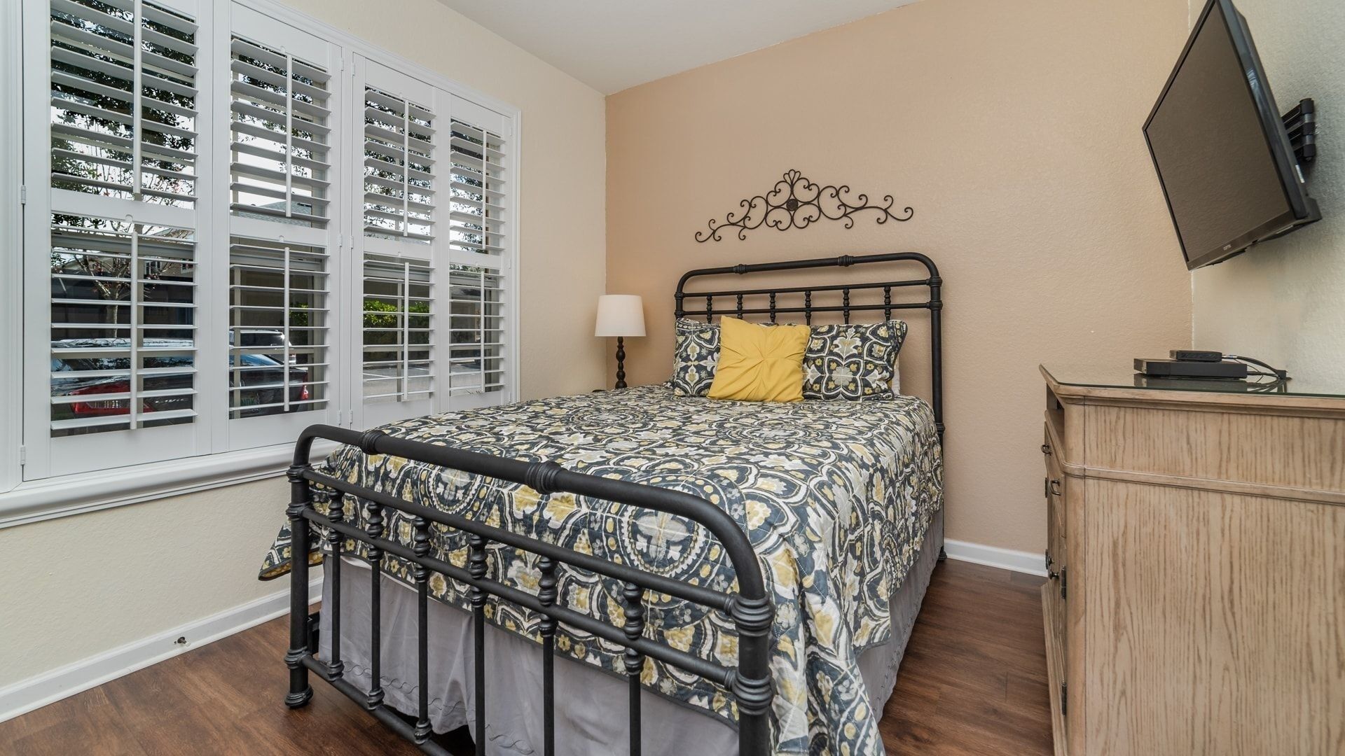 A bedroom with a bed, a nightstand, a dresser, a TV mounted on the wall, and a window with blinds.