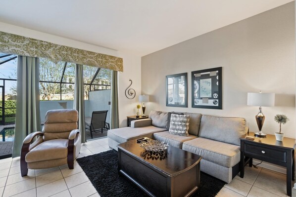 A living room with a sofa, armchair, coffee table, and a lamp. There are two framed pictures on the wall and a window with curtains. - Windsor Hills Resort 3 Bedroom Private Pool Townhome 3 Townhouse (Kissimmee)