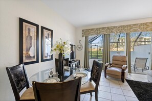A dining area with a glass table, chairs, a vase with flowers, framed pictures on the wall, and a view of the outside through large windows. - Windsor Hills Resort 3 Bedroom Private Pool Townhome 3 Townhouse (Kissimmee)