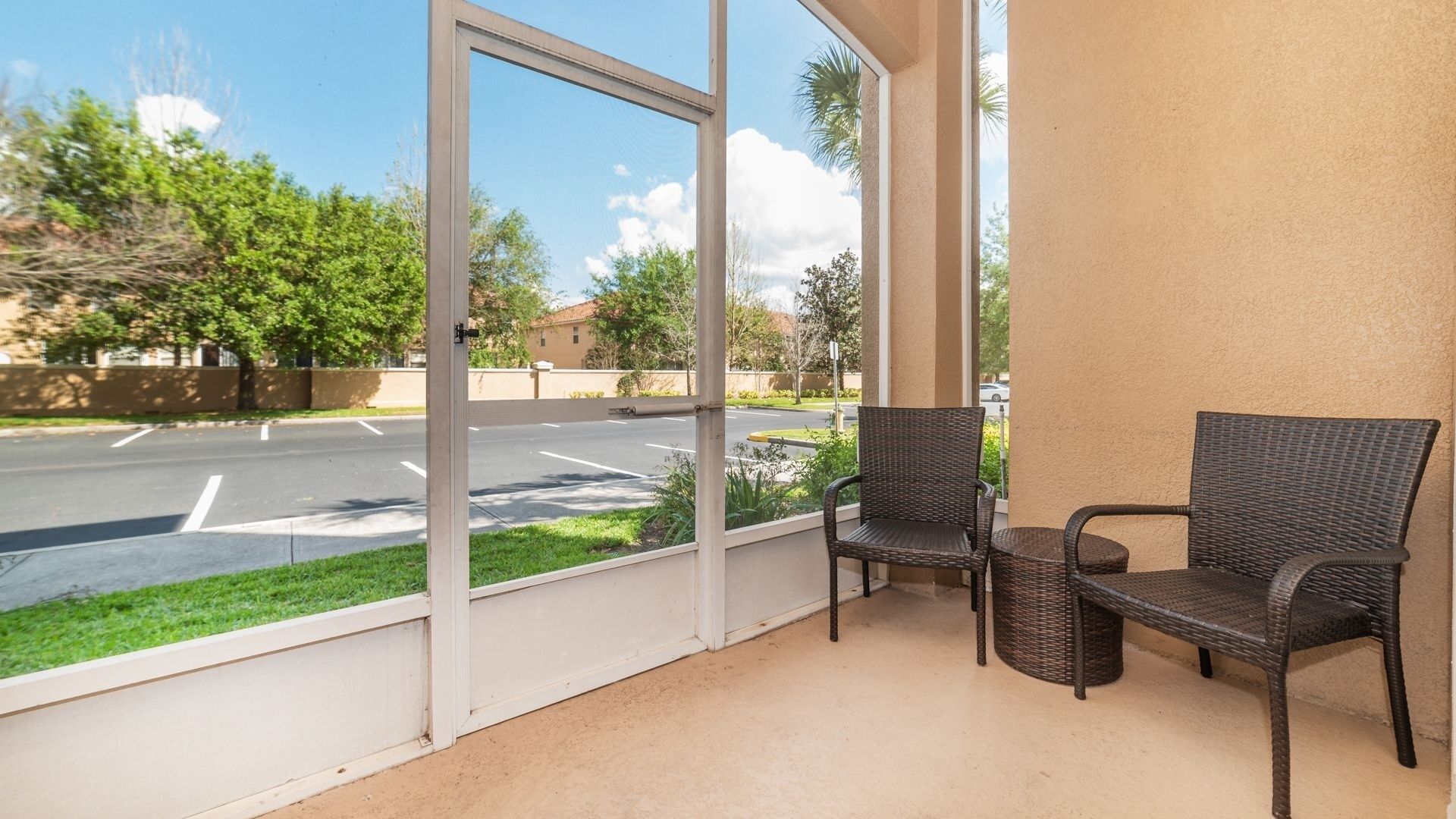 A balcony with two wicker chairs, a view of a parking lot, and a building with a beige wall.