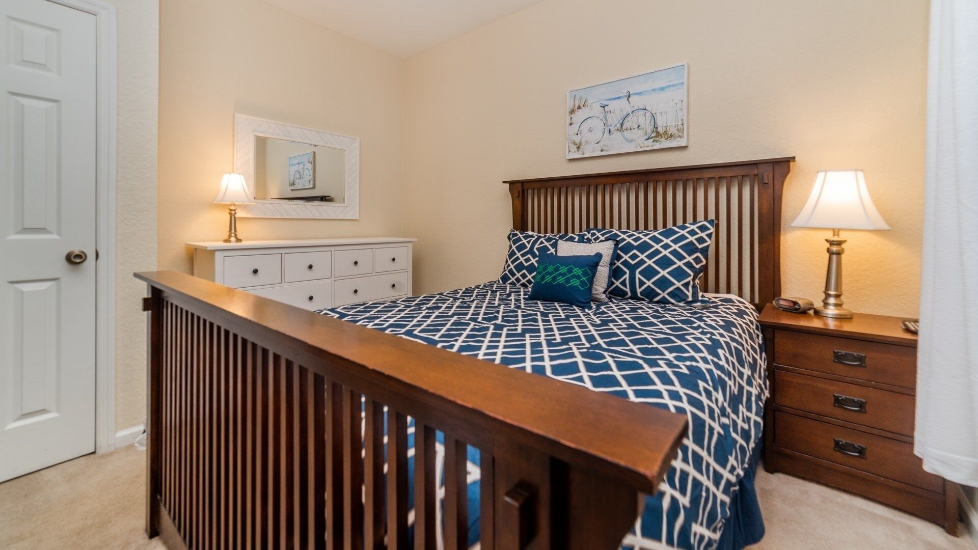 A bedroom with a wooden headboard, a bed with patterned bedding, a nightstand with a lamp, a dresser, and a door.