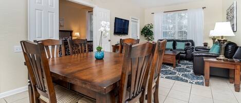 A dining area with a wooden table and chairs, a TV mounted on the wall, and a living room with a sofa and a lamp.