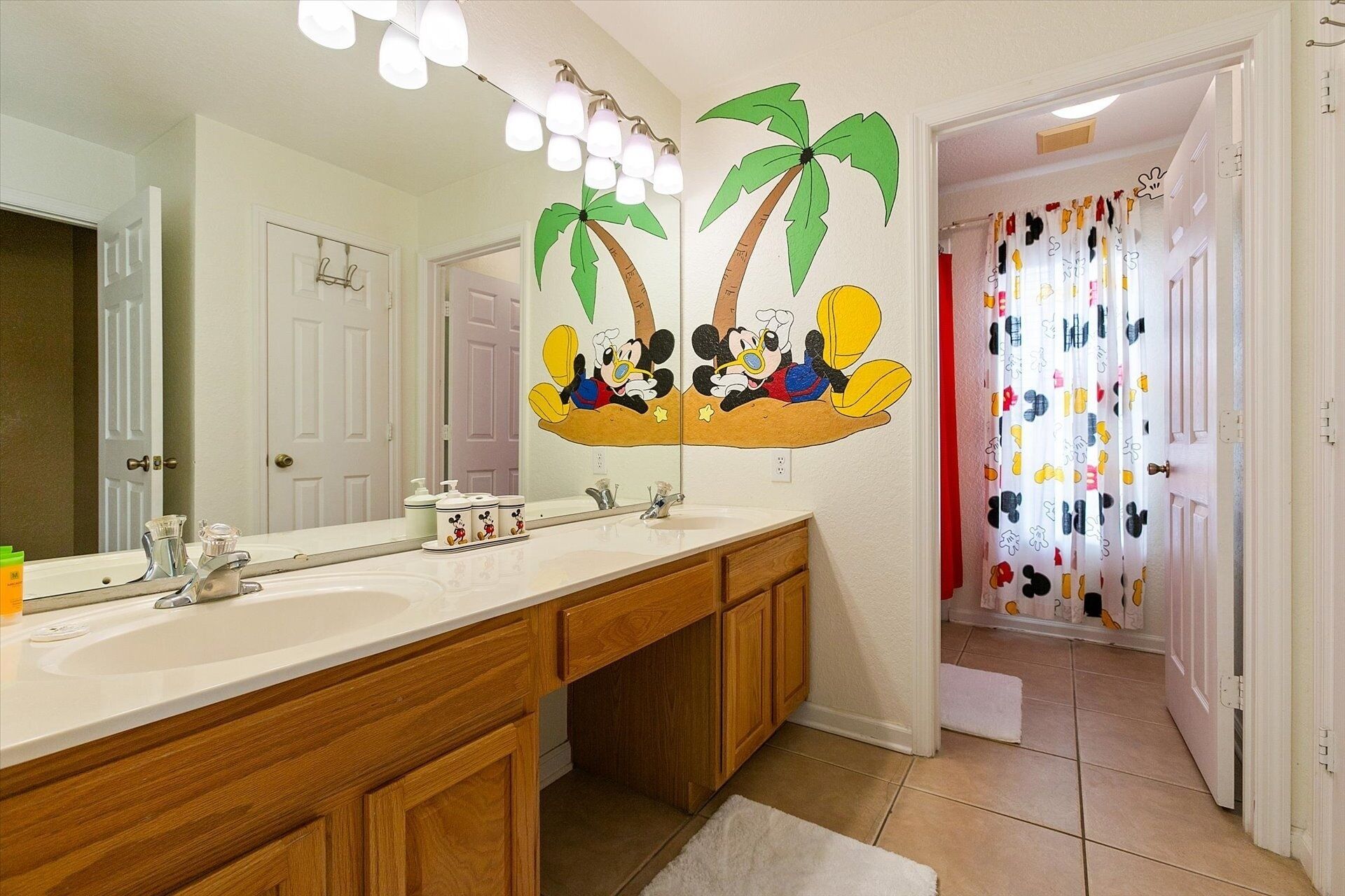 Une salle de bain avec un grand miroir, deux lavabos, des meubles en bois et une fresque murale représentant Mickey Mouse.