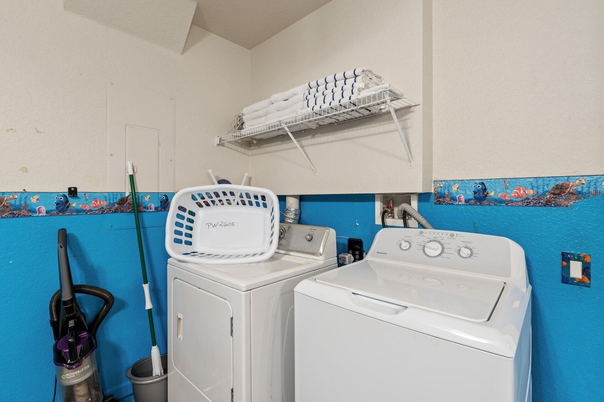 A laundry area with a front-loading washer and dryer, a white towel rack above the dryer, and a blue wall with an underwater mural.