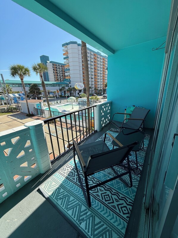 A balcony with a patterned floor, two black chairs, and a view of a pool and buildings. - Beachside Beauty Myrtle Beach studio Pool View, Lazy River, AC - Steps to Beach (Myrtle Beach)