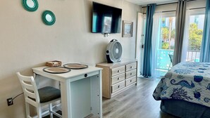 Une chambre à coucher avec un lit, un bureau et une chaise, un téléviseur, un ventilateur et une fenêtre avec des rideaux.