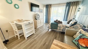 Un espace de vie compact comprenant un coin repas, un lit avec une couvre-lit à motifs, une petite table en bois et un ventilateur.