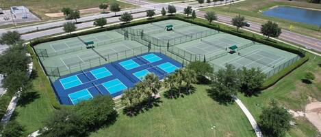 An aerial view of a tennis court complex with multiple courts, surrounded by greenery and residential buildings.