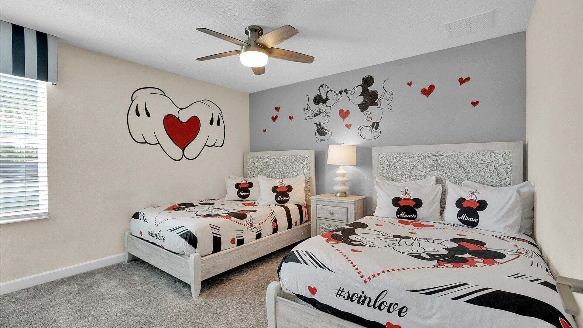 Ein Schlafzimmer mit zwei Betten, jeweils mit Bettwäsche im Minnie Maus Stil. Es gibt einen Deckenventilator, einen Nachttisch mit Lampe und eine Wand mit Herz- und Minnie Maus-Dekorationen.