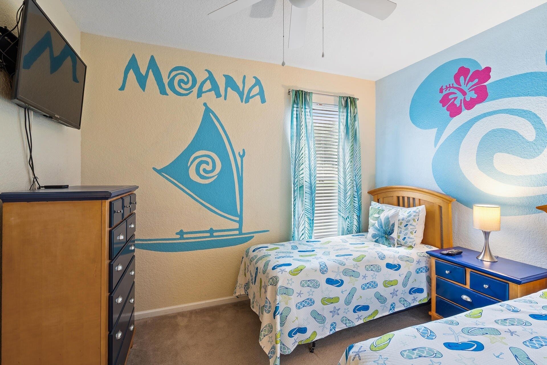 A bedroom with a sailboat mural, a bed with a tropical print, a dresser, and a TV mounted on the wall.