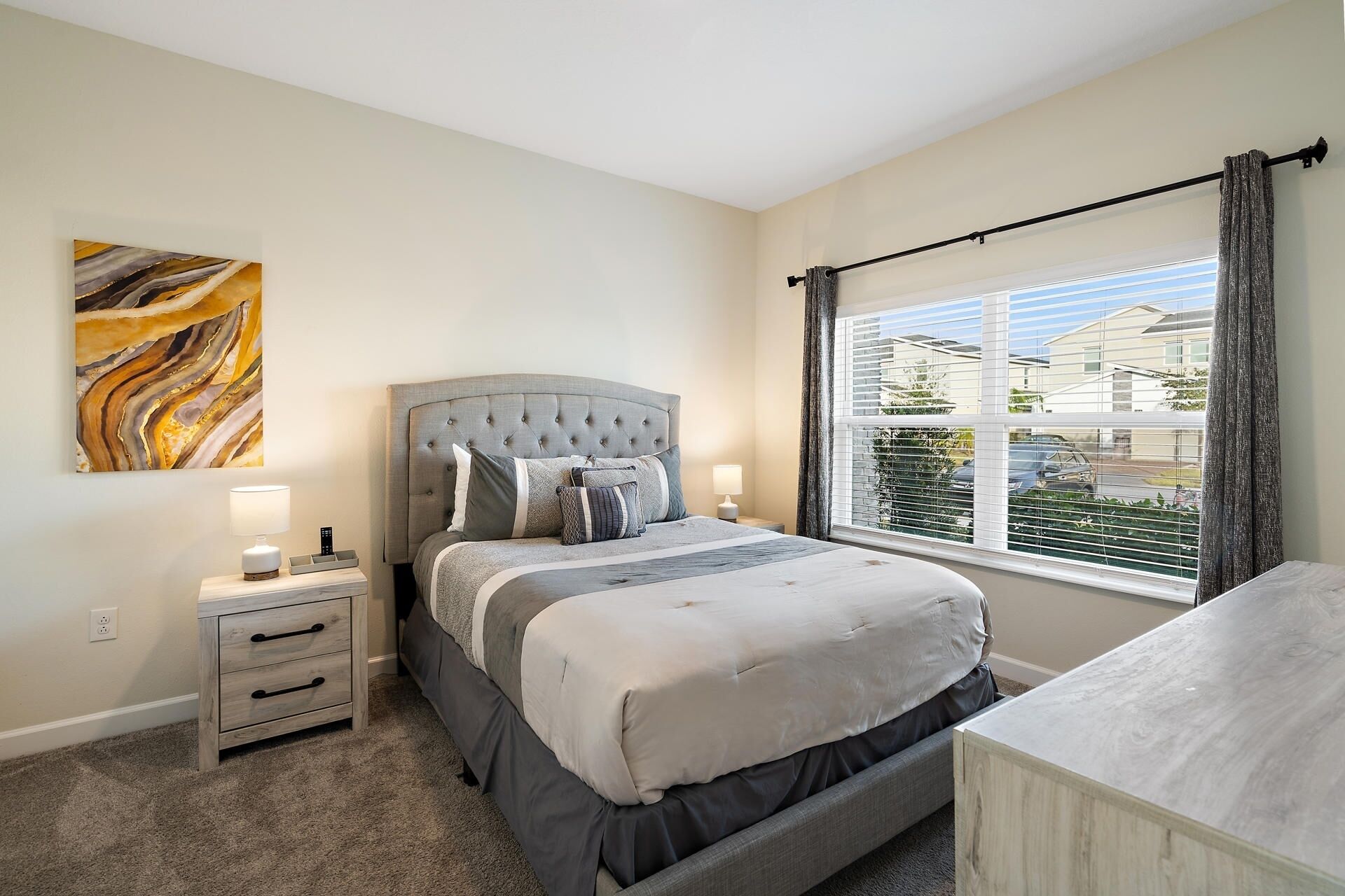 A bedroom with a tufted headboard bed, bedside tables with lamps, a large window with blinds, and a marble artwork on the wall.