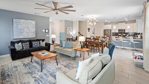 Un salon moderne avec un canapé gris foncé, un canapé beige clair et une table basse en bois. On y trouve également un coin repas avec une table et des chaises en bois, ainsi qu’une cuisine avec des meubles blancs et un coin bar.