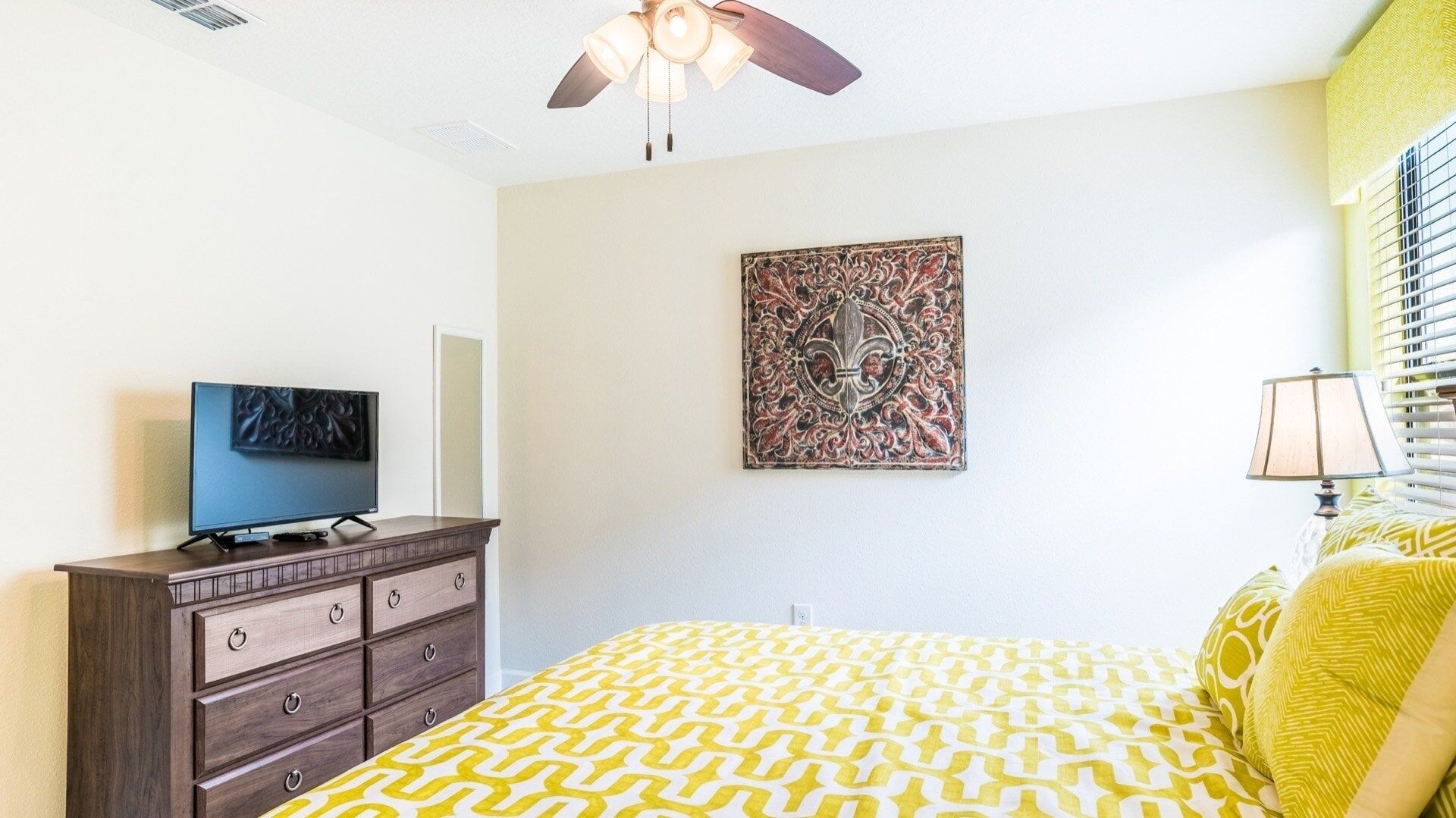 Une chambre à coucher comprenant un lit, une commode, un téléviseur, un ventilateur de plafond, une œuvre d’art accrochée au mur et une fenêtre avec des stores.