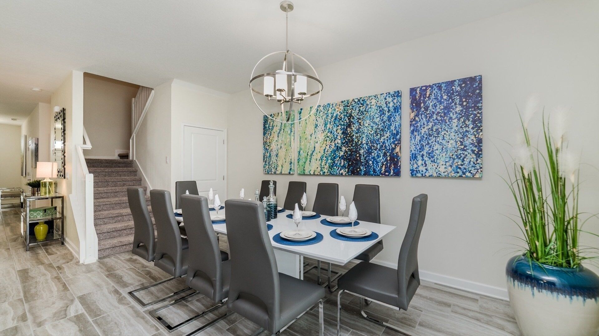 A dining area with a rectangular table set for six, surrounded by six chairs. There is a staircase leading to another level, and a hallway with a lamp and a small table with decorative items.