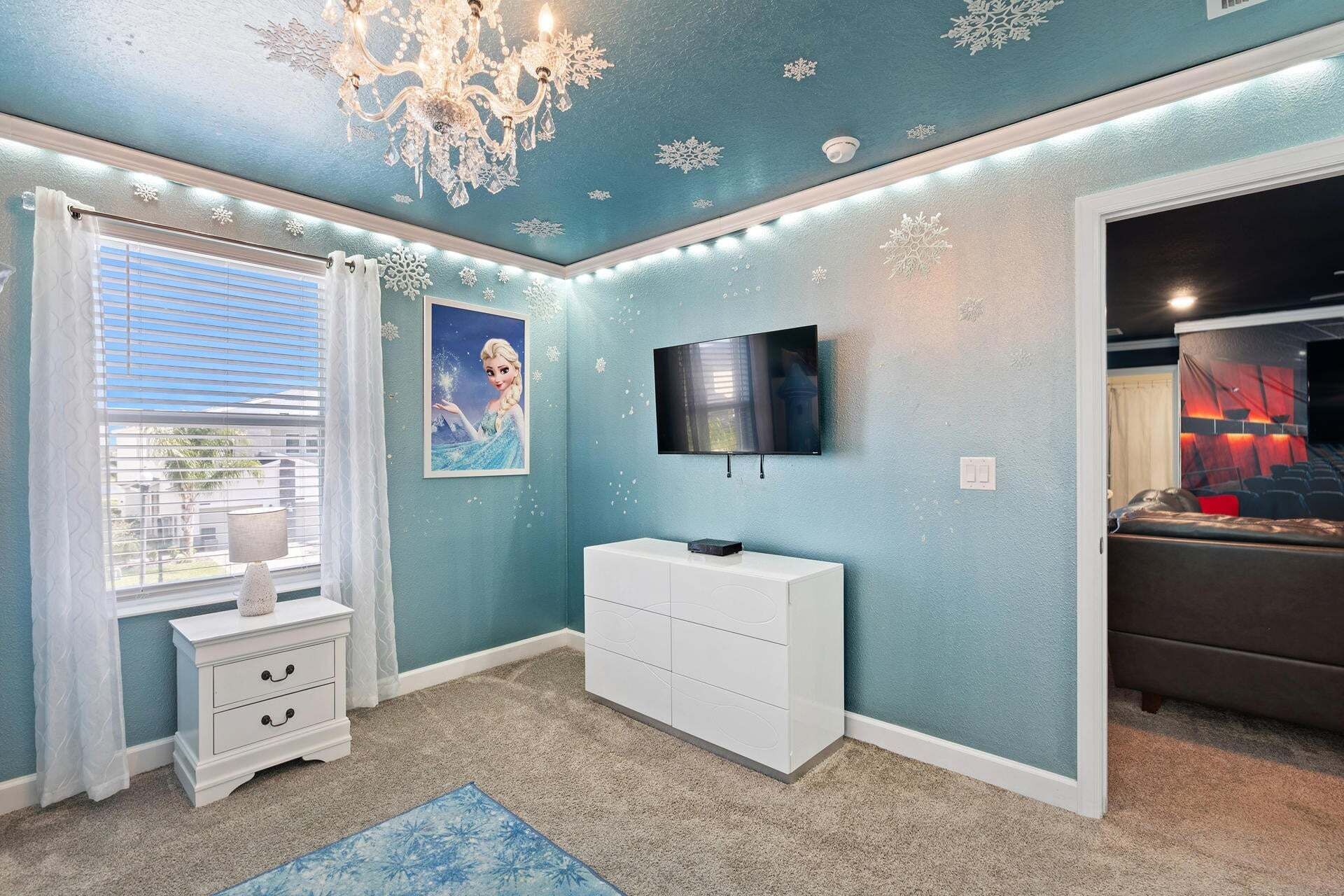 A room with a blue wall, a chandelier, a flat-screen TV, a white dresser, and a window with blinds.