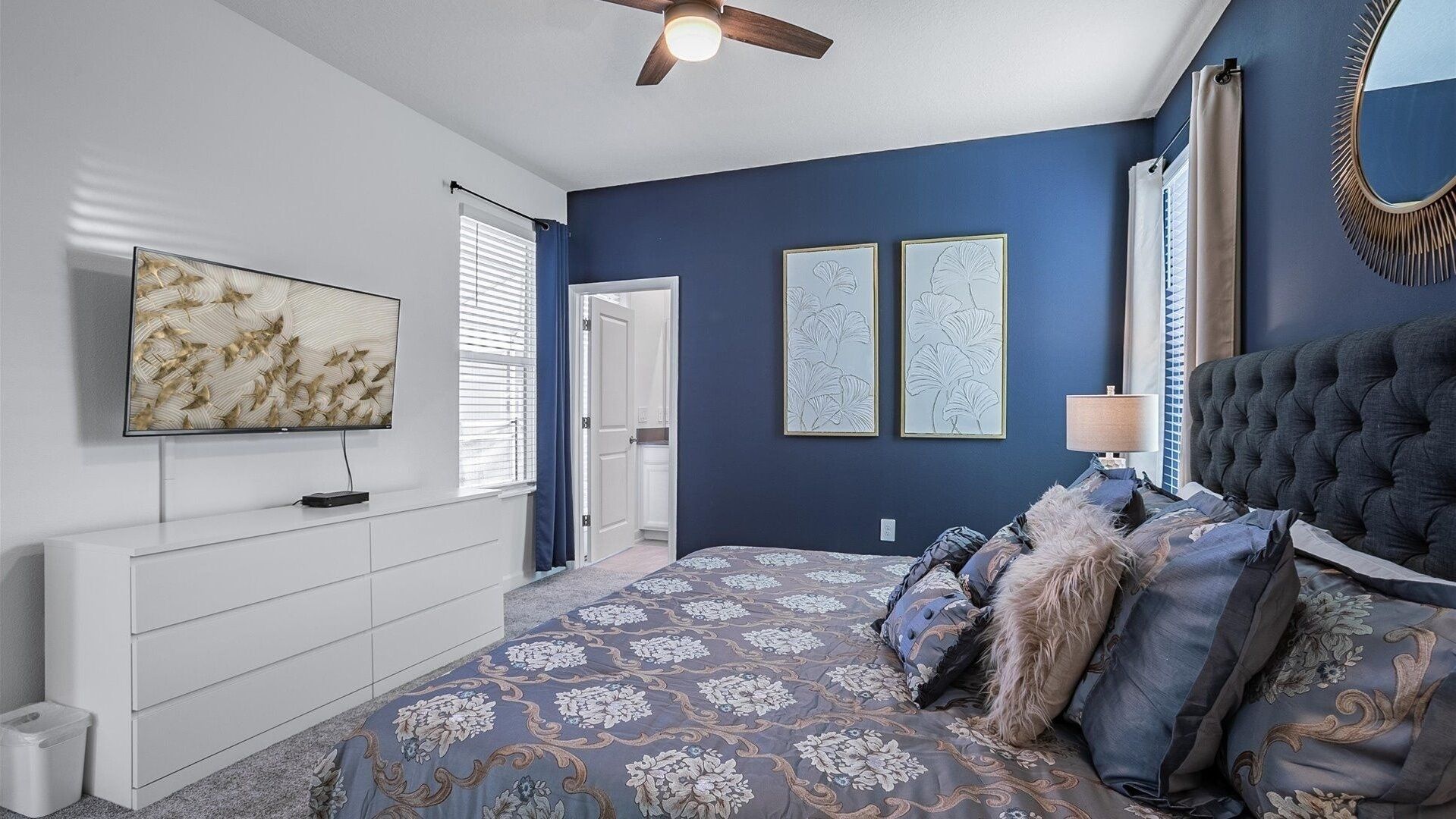 A bedroom with a large bed, a ceiling fan, a TV, and artwork on the walls.