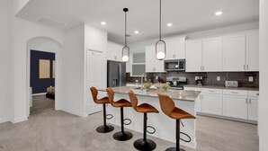 A modern kitchen with white cabinets, a central island with bar stools, and a dining area with a dark-colored wall and a door. - Championsgate Resort 5 Bedroom Private Pool Home 5 Home (Davenport)