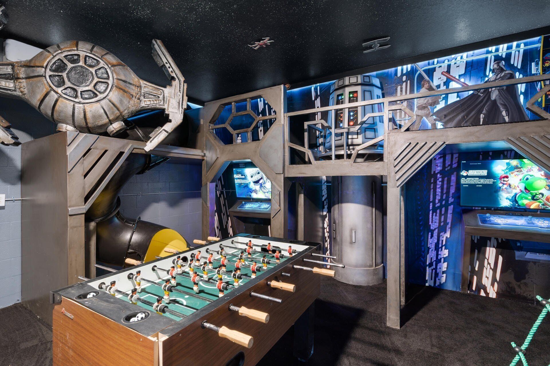 A room with a foosball table, Star Wars-themed decor, and multiple screens displaying video games.