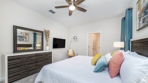 Une chambre à coucher comprenant un lit, une commode, un téléviseur, un ventilateur de plafond et une porte donnant accès à une salle de bain.