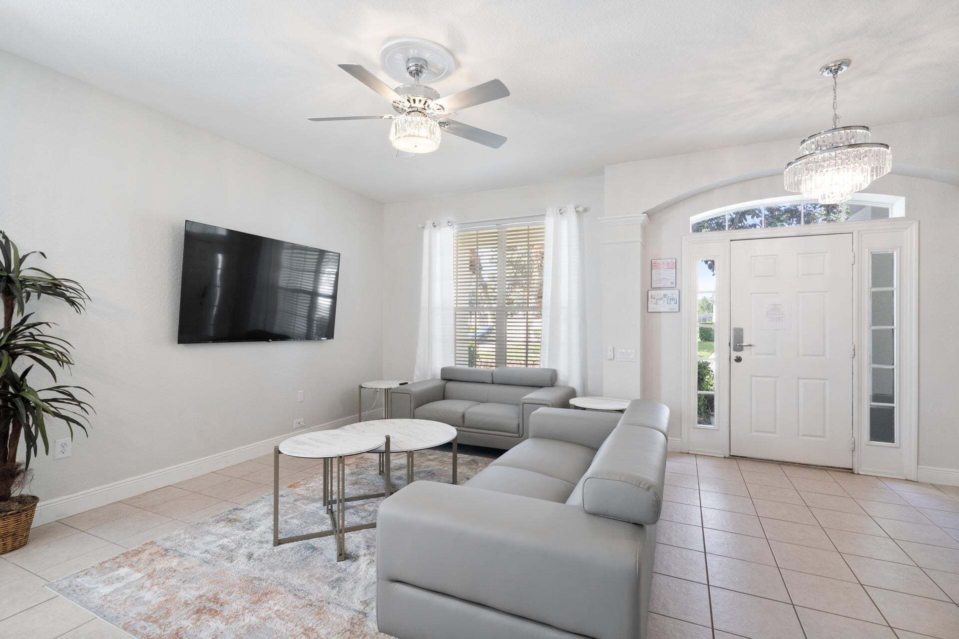 A living room with a grey sofa, a small round table, a TV mounted on the wall, a ceiling fan, and a chandelier.