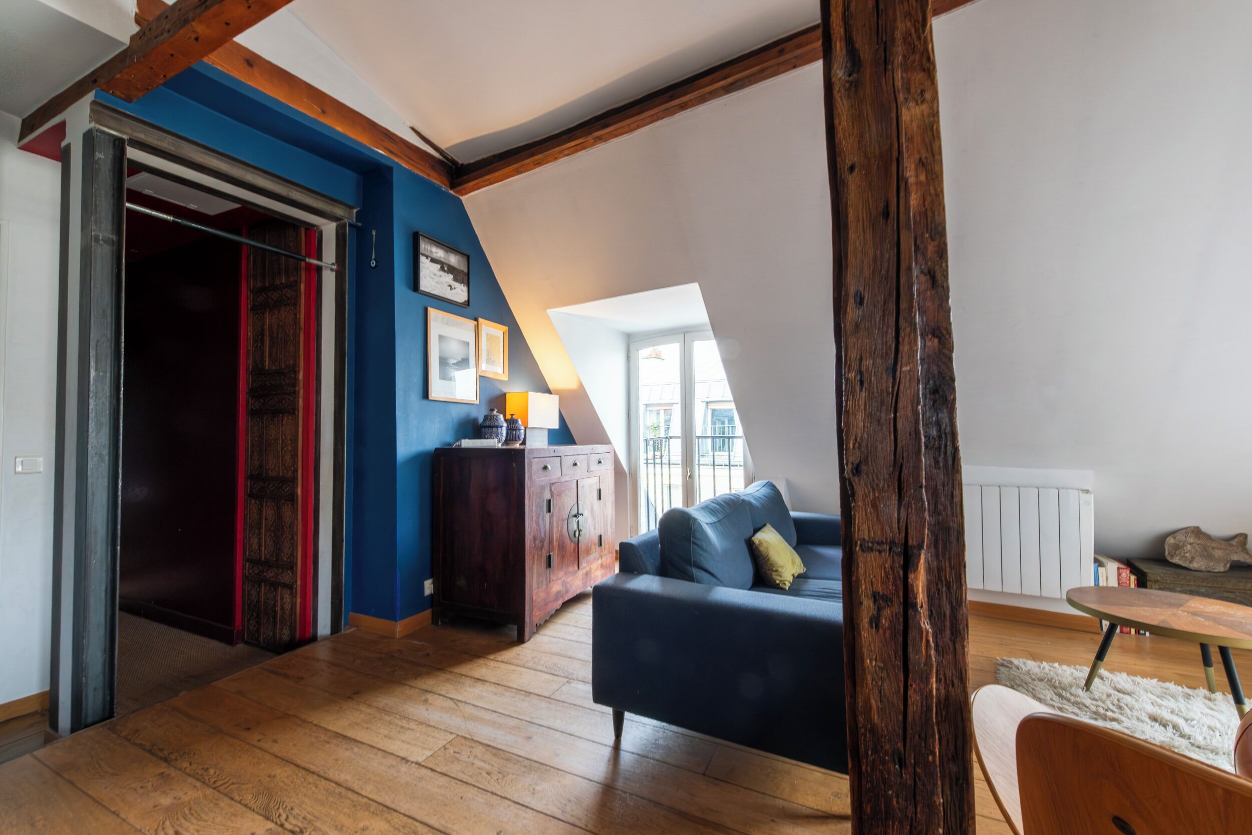 Een loft-achtige kamer met zichtbare houten balken, een blauwe accentmuur, een donkerblauwe bank en een houten vloer.