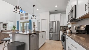 A modern kitchen with stainless steel appliances, a central island, and a view to the living area. - Championsgate Resort 6 Bedroom Private Pool+Spa Home 5 Home (Davenport)