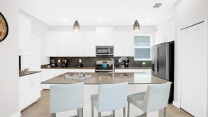 A modern kitchen with white cabinetry, a central island with bar stools, a built-in microwave, and a stainless steel refrigerator. - Championsgate Resort 5 Bedroom Private Pool Home 5 Home (Kissimmee)