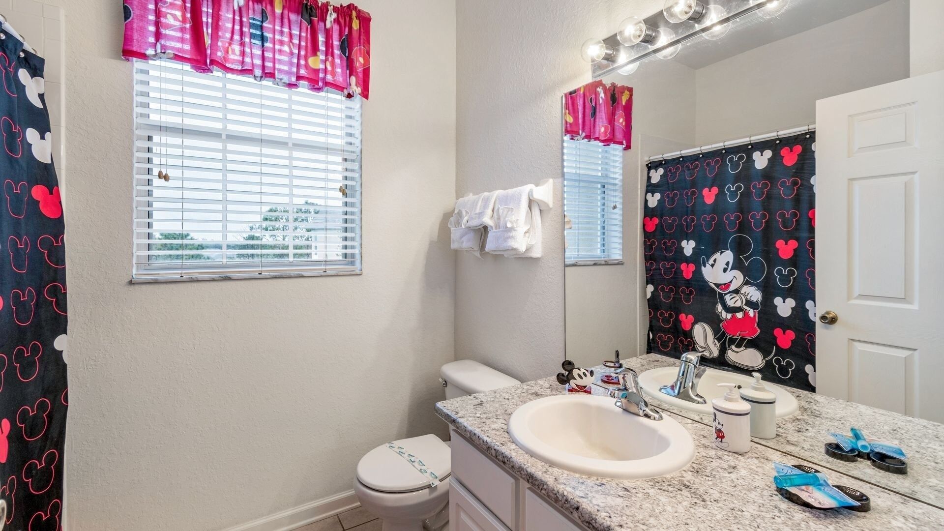 A bathroom with a toilet, sink, and mirror. There are Mickey Mouse themed curtains and a shower curtain.