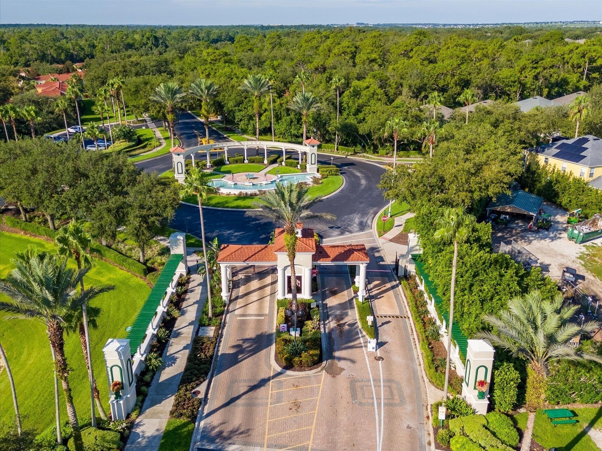 An aerial view of a gated community with a central roundabout, palm trees, and a parking area.