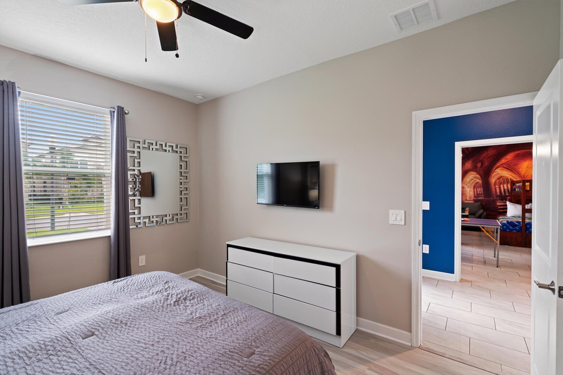 A bedroom with a bed, a TV, a ceiling fan, and a door leading to another room.