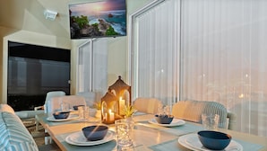 Un coin repas avec un téléviseur fixé au mur, une table dressée avec des bols et des verres, et une grande fenêtre équipée de stores.