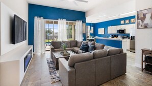 A modern living room with a grey sofa, a coffee table, and a TV. There's a kitchen area with white cabinets and a microwave. - Windsor Hills Resort 5 Bedroom Home with Game Room 5 Home (Kissimmee)