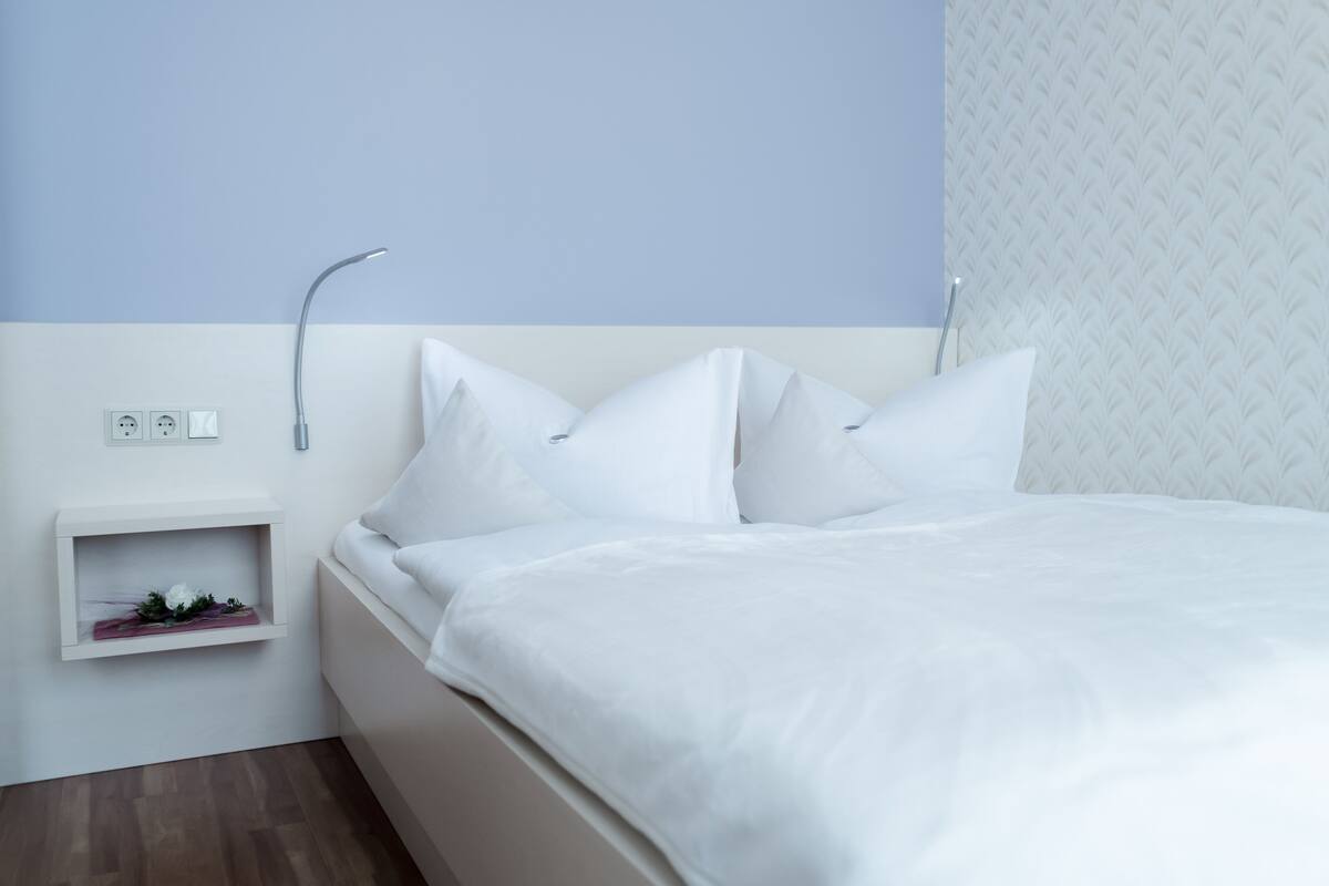 A neatly made bed with white linens and pillows, a bedside table with a small plant, and a wall-mounted light fixture.