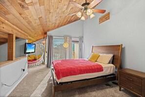 Ein Schlafzimmer mit Holzboden, einem Bett mit roter Decke, einem Nachttisch aus Holz und einem Fernseher auf einer weißen Kommode.