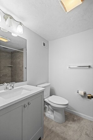 Ein Badezimmer mit weißem Waschbecken, Toilette und Spiegel. An der Wand befindet sich ein Handtuchhalter und eine Leuchte über dem Spiegel.