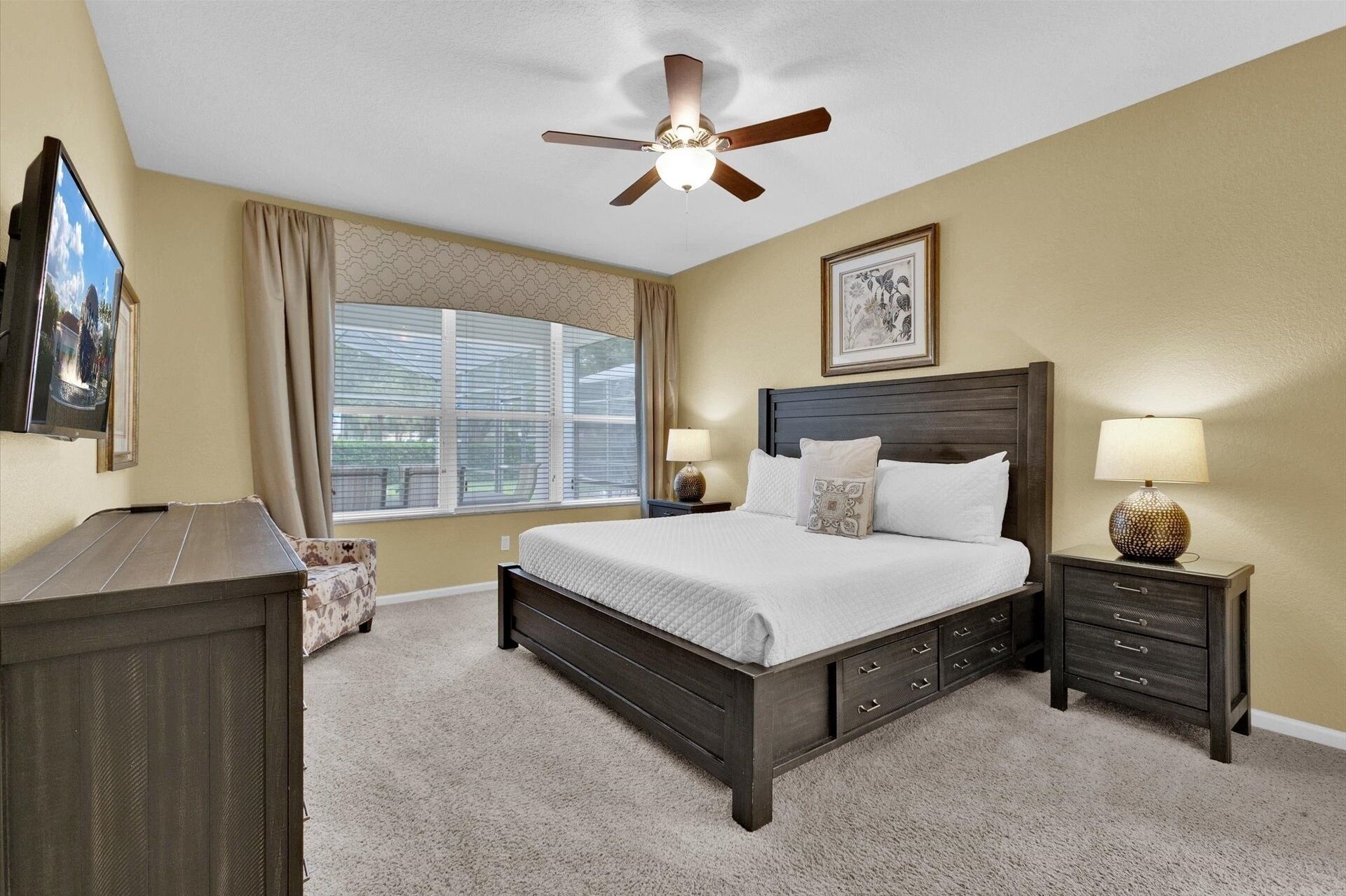 A bedroom with a bed, two bedside tables, a ceiling fan, a window with blinds, and a framed picture on the wall.
