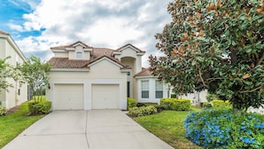 A two-story house with a tiled roof, two garage doors, and a well-maintained front yard with bushes and a tree. - Windsor Hills Resort 5 Bedroom Private Pool+Spa Home 5 Home (Kissimmee)