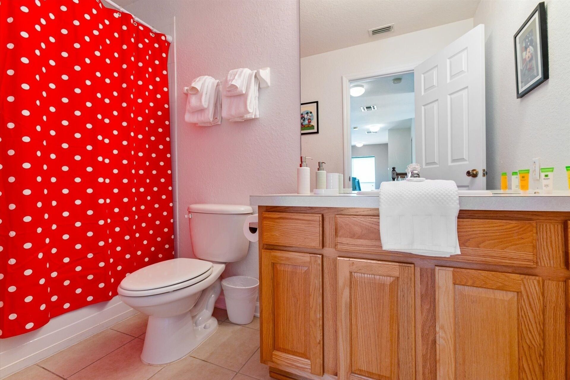 Ein Badezimmer mit dunkelrotem Duschvorhang mit weißen Punkten, einer weißen Toilette, einem Holzbadezimmer-Waschtisch mit Spiegel und einem Waschbecken mit Pflegeprodukten.