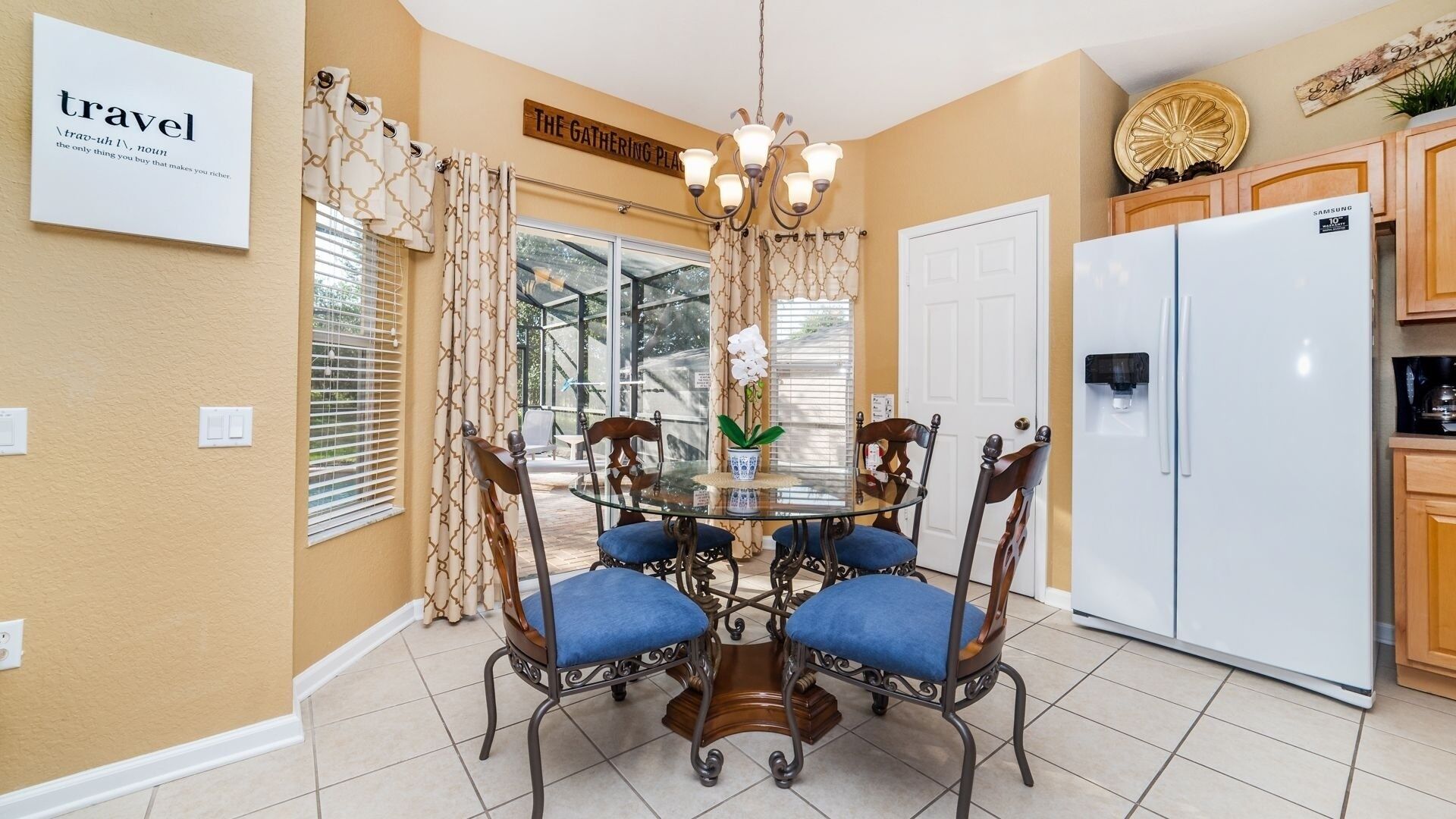 A dining area with a round glass table surrounded by four chairs, a chandelier, and a view of the outdoors through large windows.