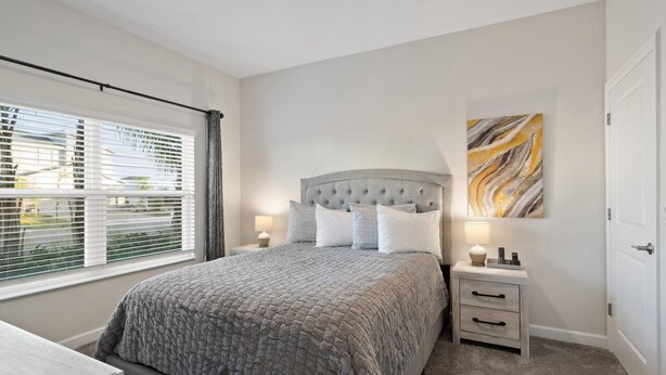 A bedroom with a bed, bedside tables, a window with blinds, a door, and a wall with a painting. - Championsgate Resort 4 Bedroom Private Pool Townhome 4 Townhouse (Davenport)