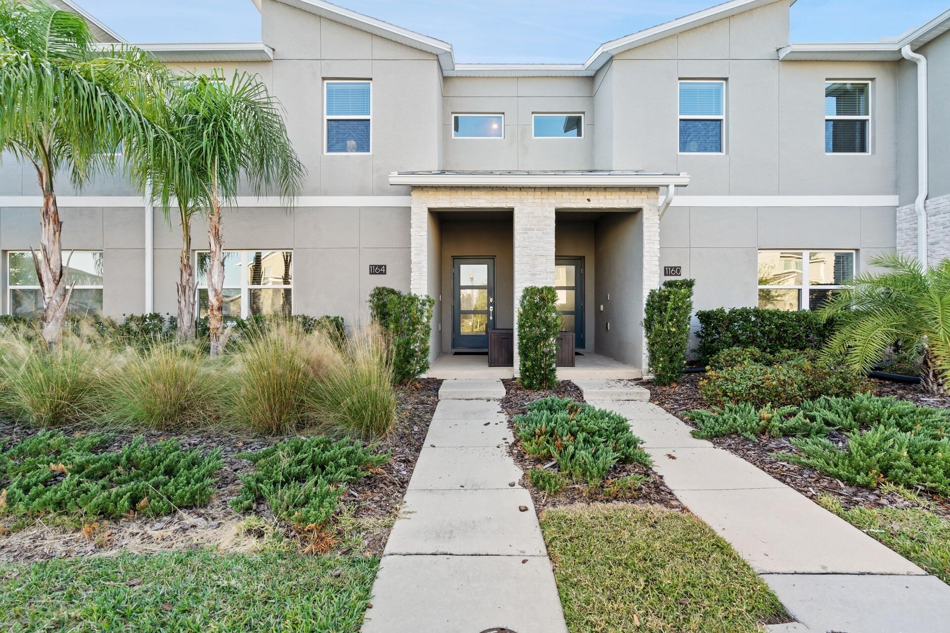 A modern two-story house with a central entrance, numbered 1164 and 1160, surrounded by a landscaped garden with various shrubs and a palm tree.