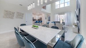 Een ruime eethoek met een lange tafel en moderne stoelen, een keuken op de achtergrond en een zithoek met een bank en gordijnen.