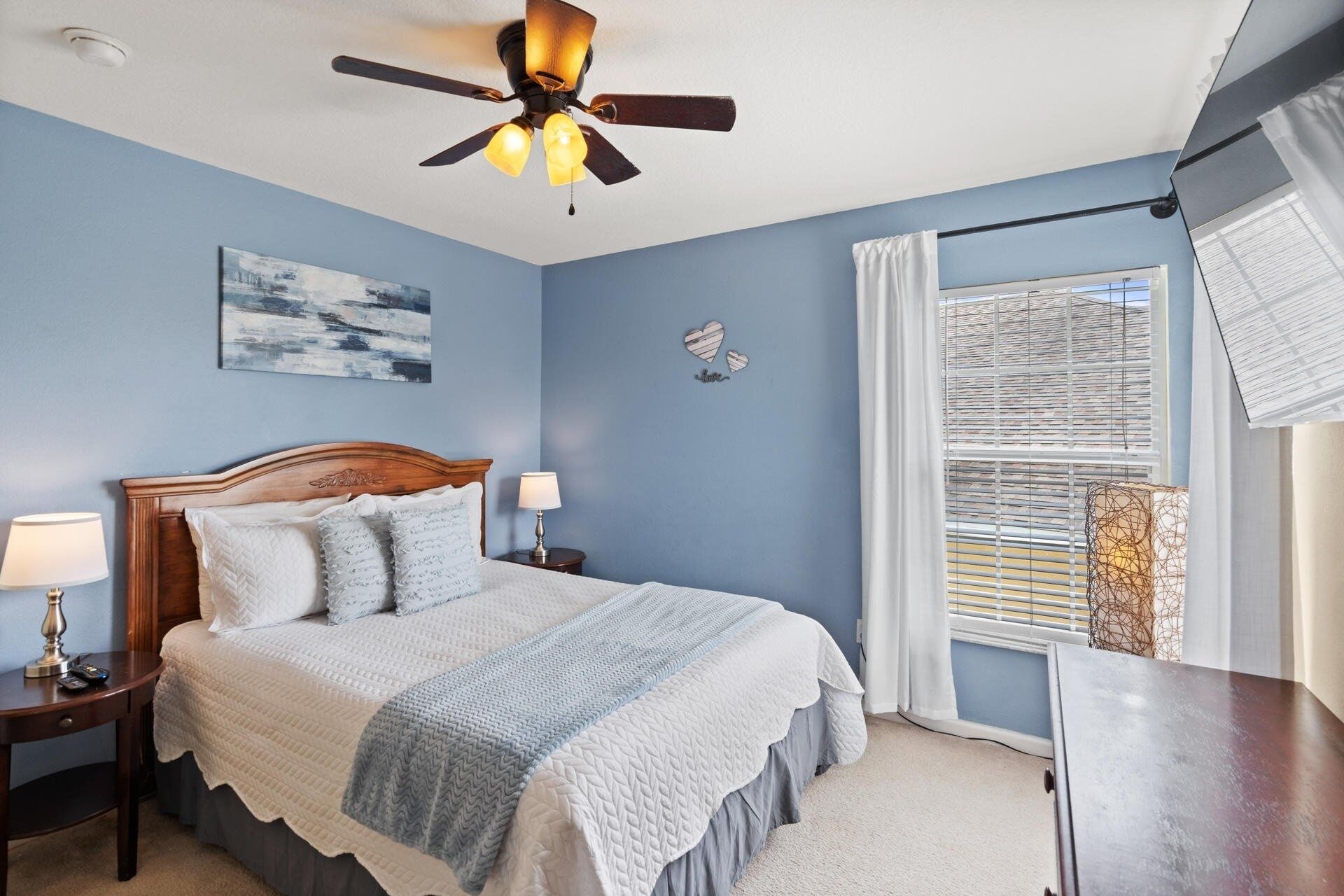 A bedroom with a bed, a ceiling fan, a nightstand, a lamp, a window with blinds, and a wall with a painting and a heart decoration.