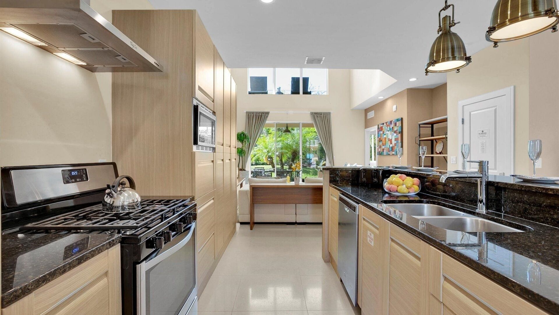 A modern kitchen with a stove, oven, and sink. There is a window with curtains, a plant, and a fruit bowl on the counter.