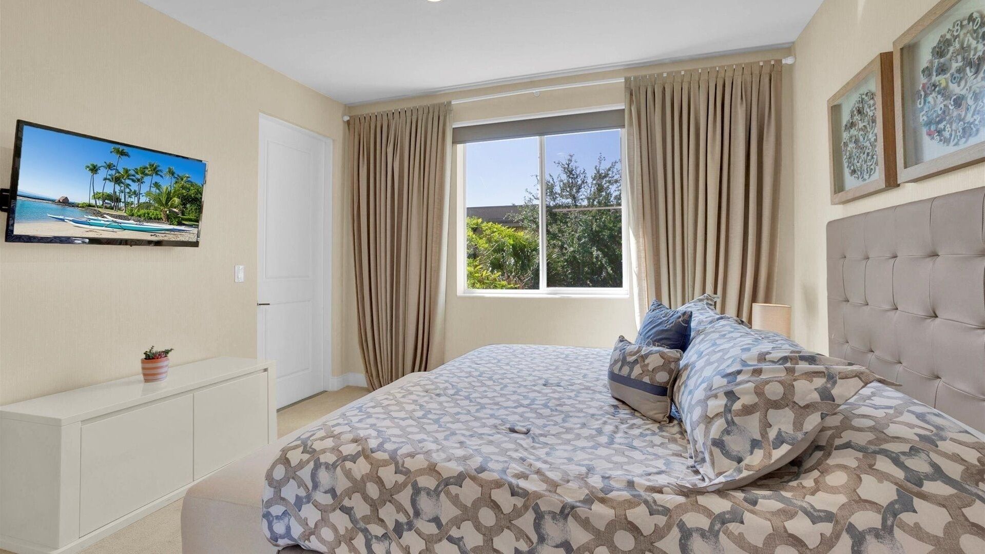 A bedroom with a bed, a television on the wall, a window with curtains, and a view of greenery outside.